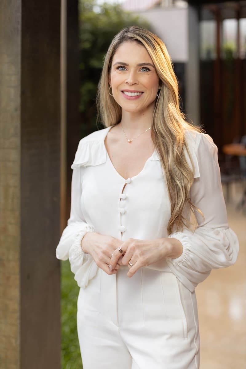 Dra. Luíse Pezzin em pé, vestindo roupa branca, em ambiente externo com estrutura de madeira
